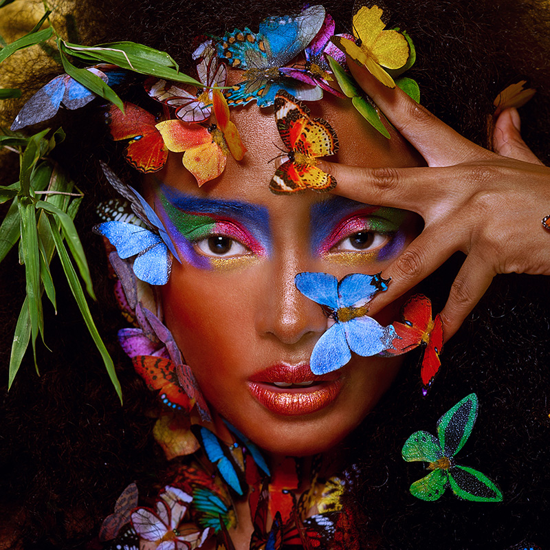 A female model with dark skin vogues with butterflies landing demurely across her face and hair.